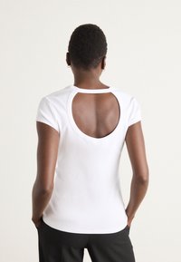T-shirt blanc ajusté avec une petite découpe ronde au dos, des manches courtes et une texture lisse, conçu pour un ajustement proche et le confort.