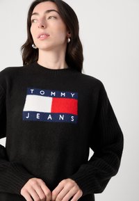 Črna jopica z rebrastimi rokavi. Ima velik, barvit pleteni logo v rdeči, beli in modri barvi z napisom "TOMMY JEANS" na prsih.