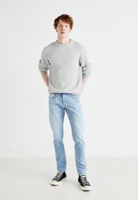 Pull gris à côtes, jeans bleu clair et baskets montantes noires. Le modèle se tient avec les mains dans les poches, fonds simple.