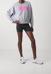 Grå DKNY sweatshirt med rosa tryck, lös passform, rund halsringning. Kombineras med svarta shorts och vita träningsskor.