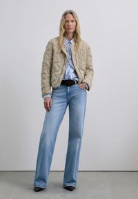 Chaqueta bomber acolchada beige, camisa de botones azul clara y jeans acampanados azul claros. Cinturón y zapatos negros. Texturas suaves, corte relajado.