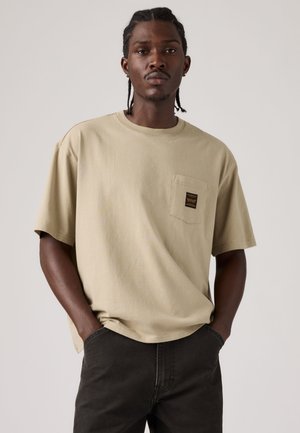 Hombre con pelo trenzado que lleva una camiseta beige de manga corta con un bolsillo en el pecho y pantalones oscuros, de pie con las manos en los bolsillos.