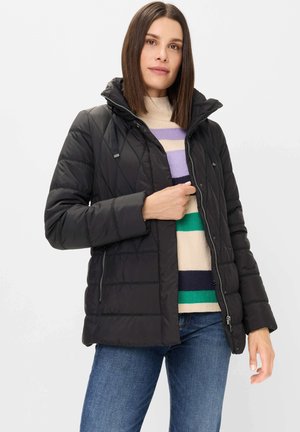 Woman wearing a black quilted jacket over a striped sweater and blue jeans, holding the jacket zipper near her chest.