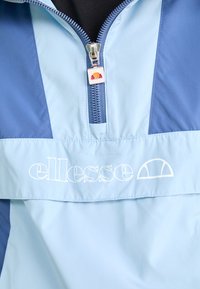 Jasnoniebieska kurtka zapinana na zamek błyskawiczny z granatowymi akcentami, z białym logo. Materiał jest gładki, z półzamkiem i wysokim kołnierzem.