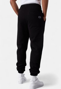 Man wearing black jogger pants and white sneakers, standing with hands in pockets, facing away against a plain white background.