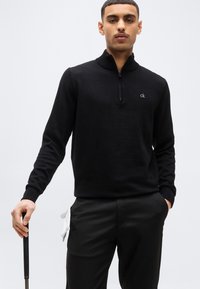 Νέος άνδρας που φοράει μαύρο πουλόβερ με φερμουάρ στο λαιμό και παντελόνι, κρατώντας ένα γκολφ ρόπαλο, με λευκά γάντια τοποθετημένα στην τσέπη του.