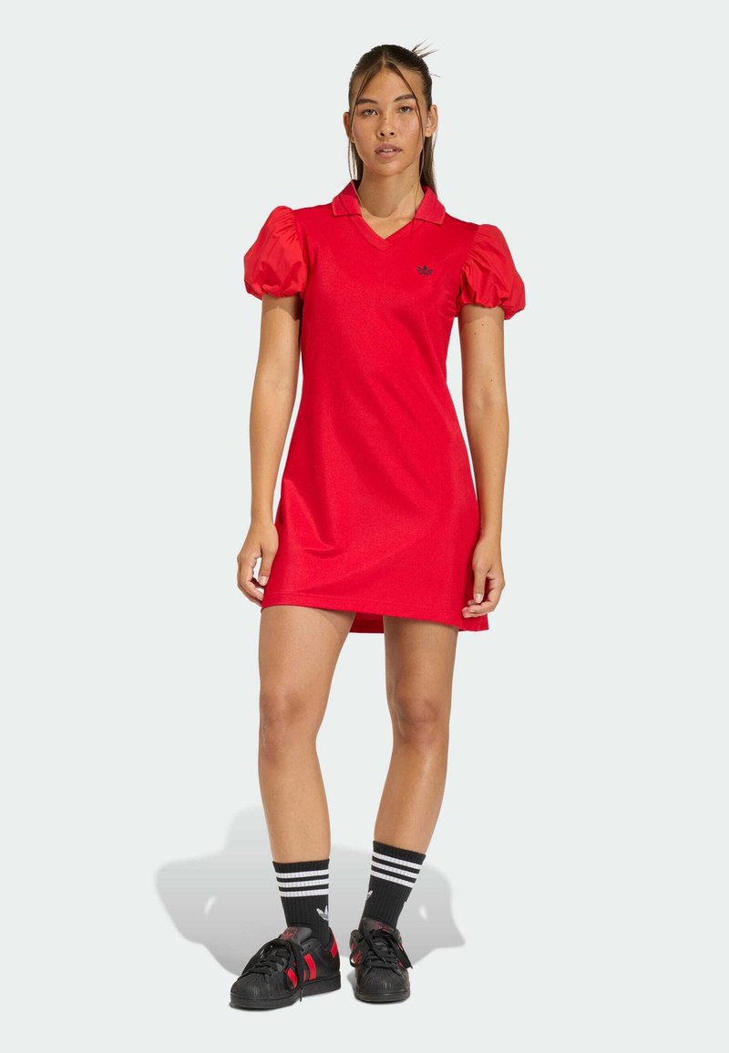 Červené šaty ve stylu polokošile s nafouklými rukávy, texturovanou látkou a vyšívaným logem. Nošeny s černými teniskami a proužkovanými ponožkami.