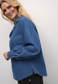 Camicia di jeans blu con dettagli arricciati, polsini abbottonati, vestibilità loose e spacco laterale. Presenta un collo a camicia e un tessuto testurizzato.