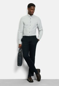 Helles graues Hemd mit Button-Down-Kragen, kombiniert mit dunklen Hosen und braunen Loafers. Hält eine dunkle lederne Aktentasche und steht vor einem weißen Hintergrund.