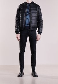 Homme portant une veste en cuir matelassé noire, un pull à motifs bleus, un pantalon slim noir et des chaussures habillées en cuir noir, se tenant contre un fond uni.