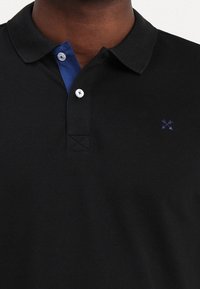 Polo noir avec un col bleu, présentant une patte de boutonnage à trois boutons et un petit logo sur la poitrine. Le tissu semble lisse et texturé.