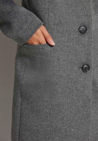 Grijze wollen jas met een zachte textuur, twee zwarte knopen en een zijzak met een hand die gedeeltelijk zichtbaar is.