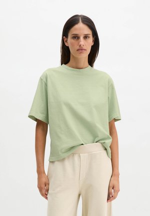 Kobieta o prostych ciemnych włosach ma na sobie luźną, jasnzięzieloną koszulkę i kremowe szerokie spodnie na tle białego tła.