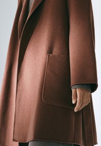Cappotto marrone di lana con un design strutturato, tasca singola sul davanti e ampi rever. Braccio mostrato che indossa un maglione grigio sotto.
