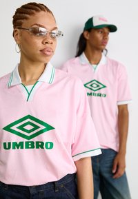 Camiseta rosa de manga curta com detalhes verdes e logo "UMBRO". Tecido texturizado, colarinho branco com acabamento verde. Jeans de ganga visíveis.