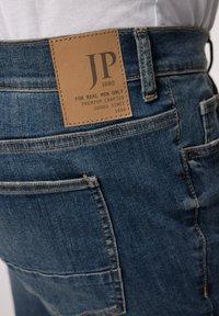 Vaqueros de mezclilla en azul oscuro con una superficie texturizada, que cuentan con una etiqueta de cuero marrón marcada "JP 1880" y bolsillos adicionales cosidos.