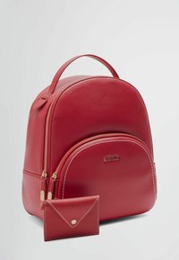 Zaino in pelle rossa a forma ovale, con cerniere dorate e una tasca frontale. Include un portacarte abbinato con chiusura a scatto e cuciture bianche.