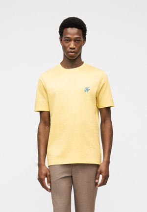 Jeune homme portant un T-shirt à manches courtes jaune pâle avec un petit logo bleu sur la poitrine et un pantalon marron clair sur un fond uni.