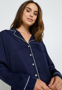 Haut de pyjama bleu marine avec passepoil blanc, manches longues, col et fermeture à boutons. Tissu doux et lisse avec une coupe décontractée.