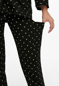 Black pants with white polka dots, showing a hand pulling a side cargo pocket open against a plain background.