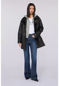 Svart läderkappa med avslappnad passform, grå huvtröja under, vit t-shirt med tryck och mörkblå utsvängda jeans. Svarta ankelstövlar.