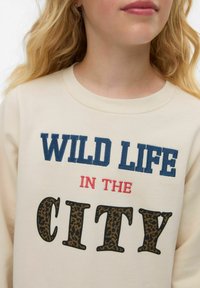 Krämfärgad sweatshirt med texten "WILD LIFE IN THE CITY" i blått, rött och leopardmönster. Mjuk tyg med rund hals och ribbade muddar.