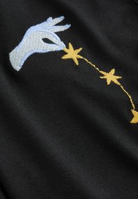 Melnā auduma virsmā ir izšūta gaiši zila roka, kas sniedzas pēc dzeltenām zvaigznēm, kas savienotas ar dzeltenu diegu, radot fantāzijas pilnu dizainu.