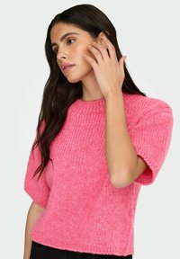 Femme aux longs cheveux foncés portant un pull tricoté à manches courtes de couleur rose vif, touchant son visage d'une main, regardant sur le côté.