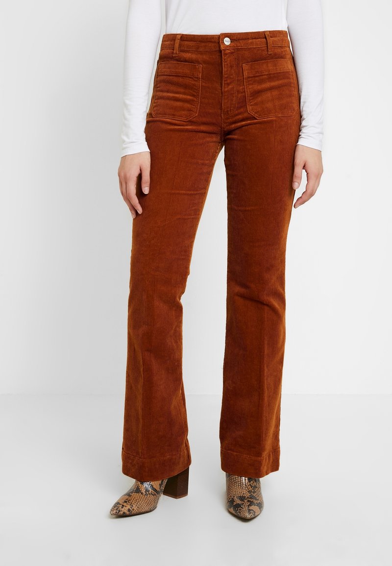 Pantalon en velours côtelé couleur rouille avec poches plaquées à l'avant, porté avec un haut blanc à manches longues et des bottines marron en peau de serpent à talons.