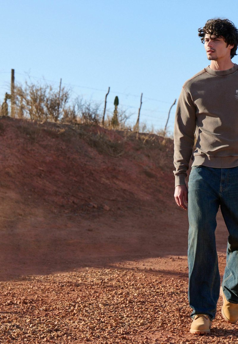 Homme aux cheveux bouclés portant un sweat-shirt marron, un jean bleu et des bottes couleur beige marchant sur un chemin de terre rocailleux près d'une clôture et d'une végétation sèche sous un ciel dégagé.
