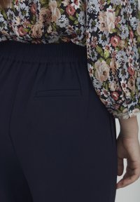 Jūrų laivyno elastingos kelnės su užpakaline kišene, derinamos su gėlėmis dekoruota bluzone, kurioje pavaizduoti įvairių spalvų rožių raštai tamsiame fone.