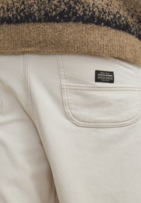 Beżowe bawełniane spodnie z dwiema tylnymi kieszeniami, widocznymi szwami oraz czarną metką z napisem "JACK & JONES" w białym kolorze.
