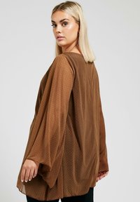 Top marrone trasparente con maniche lunghe, presenta piccoli pois neri. Vestibilità ampia con scollatura rotonda, leggera trasparenza e texture liscia.