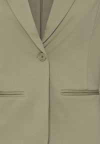 Gros plan d'un blazer beige avec un col à revers cranté, une fermeture par un bouton et deux poches passepoilées horizontales.