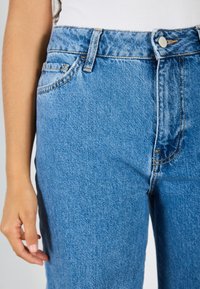 Niebieskie jeansy denimowe z wysokim stanem, z pięcioma kieszeniami, mosiężnymi detalami i przeszyciami na pasku oraz szwach.