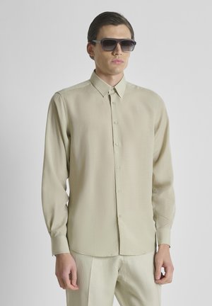 Čovjek u bež dugoj košulji s gumbima, odgovarajućim hlačama i velikim crnim sunčanim naočalama na jednostavnoj svijetloj pozadini.