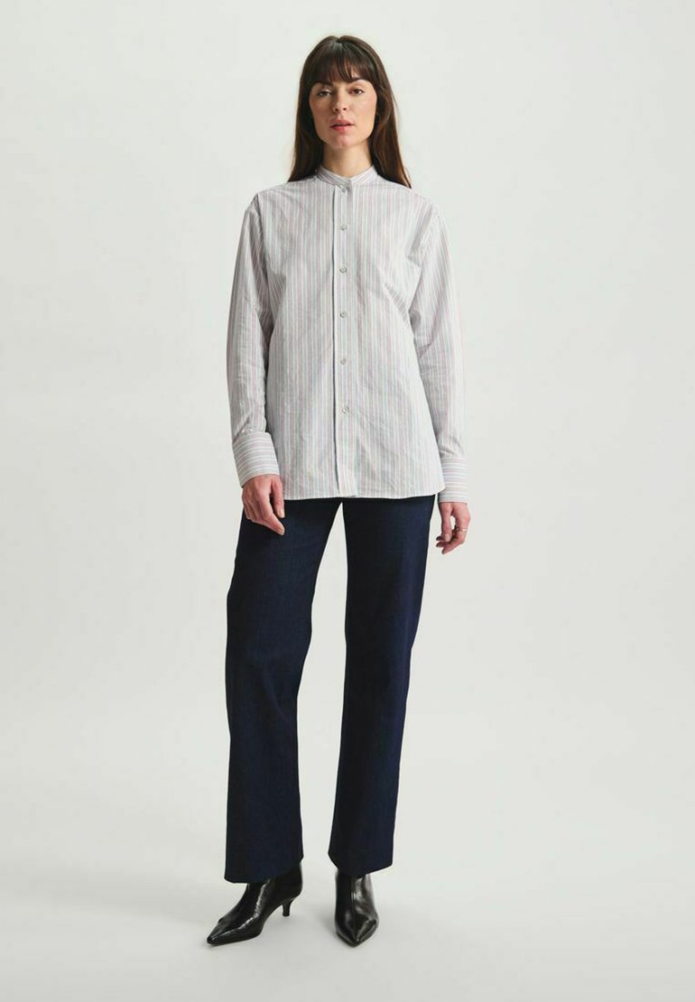 Chemise blanche à boutons avec des fines rayures multicolores, manches longues et col mandarin, assortie à un pantalon droit marine foncé et des bottines à talons noires.