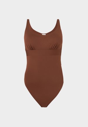 Žena stojaca s pravou rukou na boku, oblečená v hnedých jednodielnych plavkách a čiernych šľapkách na svetlom jednoduchom pozadí.