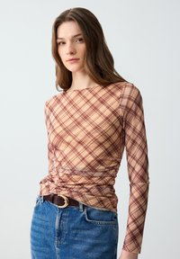 Lange mouwen geruite top in beige en bordeauxrood, met geplooide zijkanten en een ronde halslijn. Gecombineerd met hooggewaisted blauwe denim jeans.