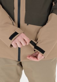 Giacca impermeabile beige e marrone con chiusura a zip, polsini in Velcro e tessuto testurizzato. Mani regolano l'apertura delle maniche.