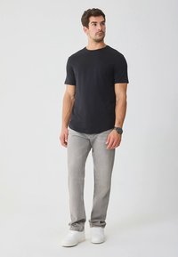 Zwart T-shirt met korte mouwen van katoen, gecombineerd met lichtgrijze spijkerbroek en witte sneakers. Model staat rechtop met een neutrale uitdrukking.