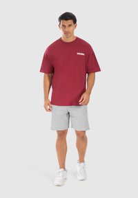 Camiseta de algodón color burdeos con mangas cortas y logo blanco, combinada con pantalones cortos gris claro y zapatillas blancas. Atuendo atlético simple y casual.
