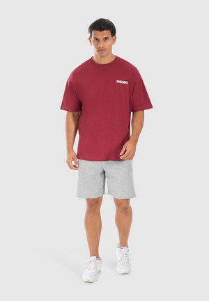 T-shirt in cotone marrone scuro con maniche corte e logo bianco, abbinata a pantaloni corti grigio chiaro e sneakers bianche. Un semplice outfit sportivo e casual.