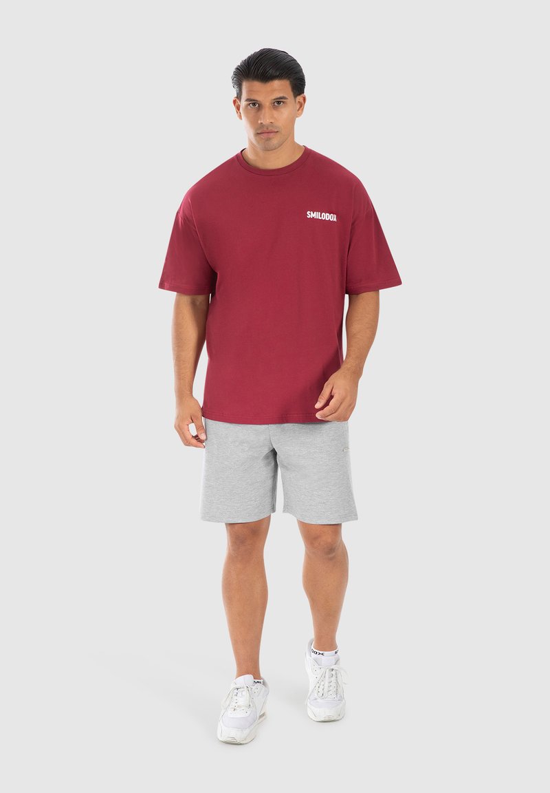 Camiseta de algodón color burdeos con mangas cortas y logo blanco, combinada con pantalones cortos gris claro y zapatillas blancas. Atuendo atlético simple y casual.