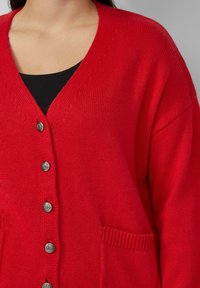 Vrouw draagt een rood gebreid vestje met knopen over een zwart topje, toont de bovenste torso en nek zonder zichtbare gezichtskenmerken.