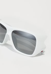 Óculos de sol brancos com armações angulares, lentes escuras e um acabamento texturizado. Apresenta bordas externas pontiagudas e discretos acentos vermelhos.