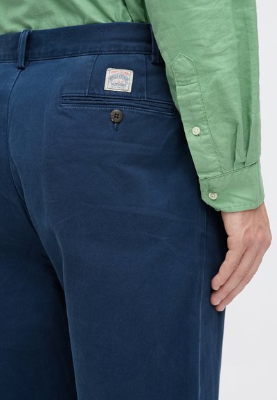 Vīrietis, tērpies tumši zilos Polo Ralph Lauren šortos un zaļā garām piedurknēm pogājamā krekla, redzams no muguras vidukļa līdz rokai.