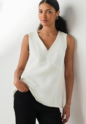 Sötét, göndör hajú nő alacsony lófarokkal, ezüst karika fülbevalót, fehér ujjatlan V-nyakú felsőt és fekete nadrágot visel, fehér háttér előtt pózolva.