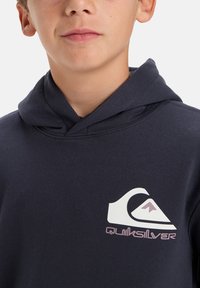 Navy blue hooded sweatshirt made of cotton, featuring a white Quiksilver logo on the chest with purple accents. Smooth texture, casual design.