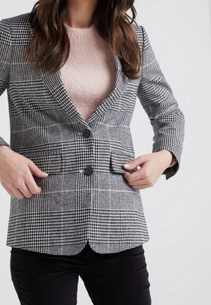 Femme ajustant un blazer à carreaux noir et blanc porté sur un pull duveteux rose clair et associé à un pantalon noir, sur un fond clair.
